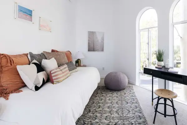 A white daybed piled with plush pillows sits in a bright room with arched windows.