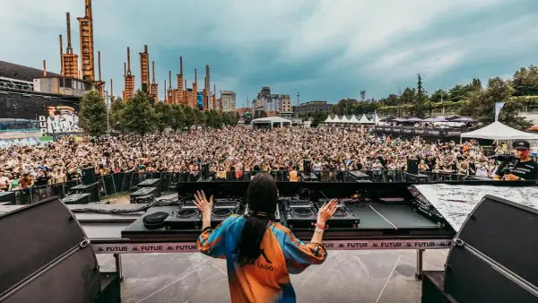 DJ set al futur festival, folla in delirio sotto un cielo nuvoloso, con un'architettura industriale sullo sfondo.