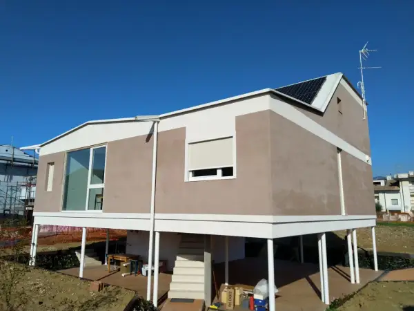 Casa moderna con intonaco argilla, pannelli solari e ampie vetrate. La struttura è rialzata su pilastri bianchi.