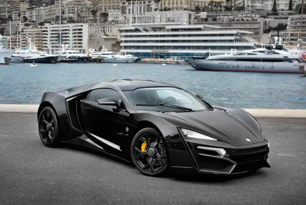 Czarny Lykan Hypersport z żółtymi zaciskami hamulcowymi na tle portu z luksusowymi jachtami i budynkami.