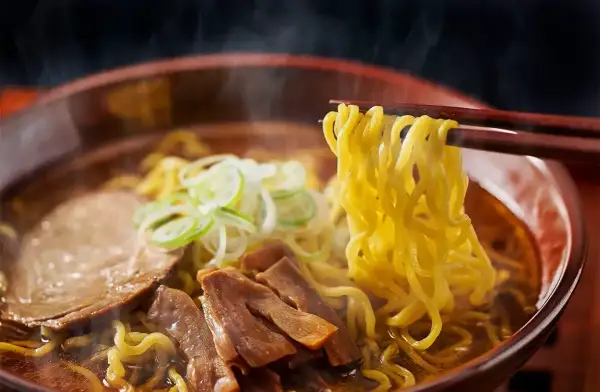 En skål med ångande ramen, med en skillnad på nudlar och pasta som tydligt syns. Nudlarna lyfts med ätpinnar, med kött och grönsaker i buljongen.