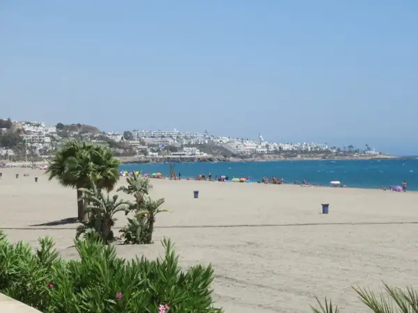 La **playa de la Mena** se extiende con arena dorada y palmeras. Al fondo, un pueblo blanco se asoma al mar azul.
