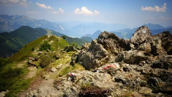 Senderos marcados con rojo y blanco serpentean por las laderas verdes de los Picos de Europa en verano, con rocas imponentes y un valle brumoso al fondo.