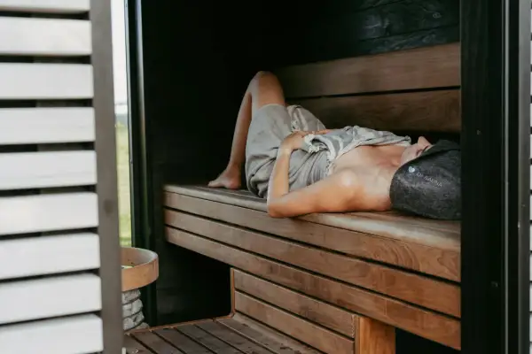 Kobieta w czepku "OF SAUNA" leży na drewnianej ławie w saunie, relaksując się. Dowiedz się, jak oddychać w saunie, by w pełni czerpać z jej dobrodziejstw.