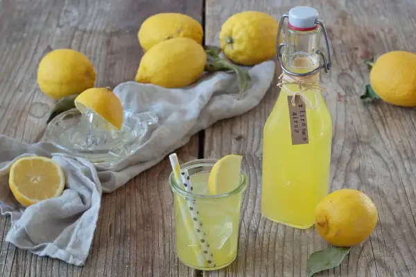 Frische Zitronen, ein Glas mit Eis und Strohhalmen, sowie eine Flasche selbstgemachter Zitronensirup auf Holzhintergrund.