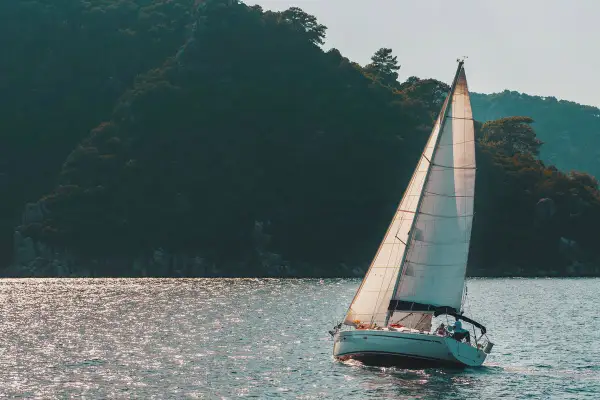 Poppa barca a vela bianca solca acque scintillanti, con una rigogliosa collina verde sullo sfondo.