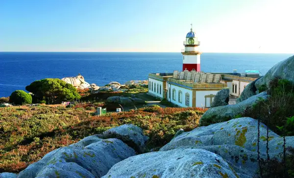 Faro en las islas Galicia, con el mar azul de fondo y rocas cubiertas de vegetación.