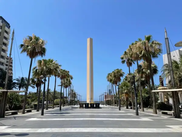 Un obelisco se alza en la plaza de las velas Almería, flanqueado por palmeras y un cielo azul despejado.