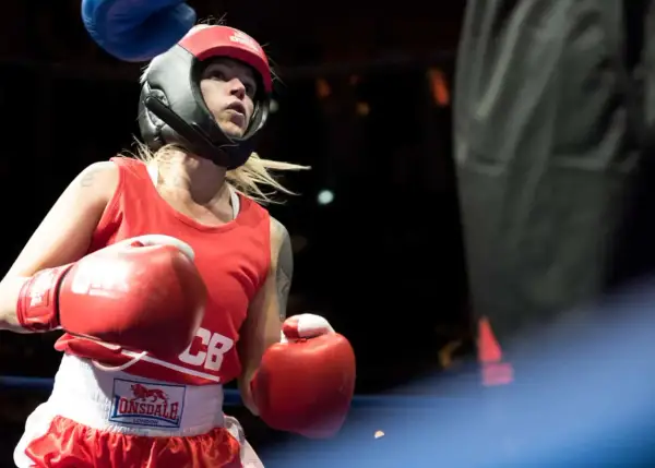 A determined female boxer in red gear and headguard prepares for a white collar boxing match, her red gloves raised.