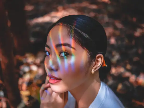 Un occhio a mandorla cattura la luce arcobaleno, riflessi colorati danzano sul suo viso mentre guarda pensierosa.