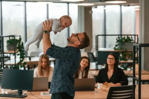 Szczęśliwy ojciec podrzuca dziecko w biurze, podczas gdy koleżanki pracują. Czy urlop wychowawczy wlicza się do stażu pracy?