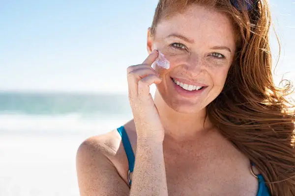 Donna sorridente con lentiggini applica crema solare sul viso in spiaggia. Si può mettere il fondotinta sopra la crema solare per un look perfetto.