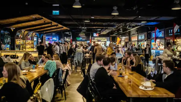 Tętniący życiem food hall Metropolia, gdzie ludzie jedzą i piją przy długich stołach, a w tle widać bary i stoiska z jedzeniem.