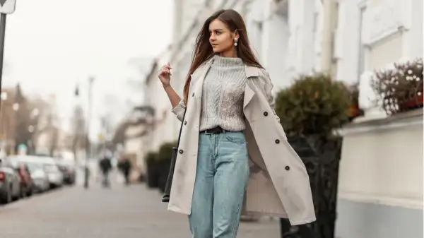 Styling Mom Jeans: Ein lässiger Look mit Strickpullover, Trenchcoat und Perlenschmuck auf der Straße.