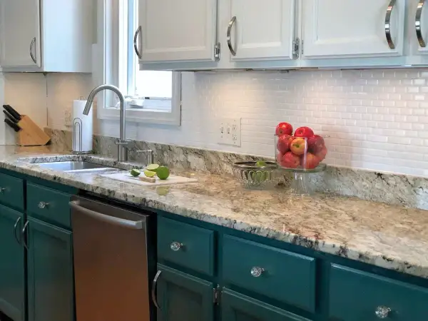 A kitchen with a white subway tile backsplash, granite countertops, and teal cabinets. A bowl of apples sits on the counter.