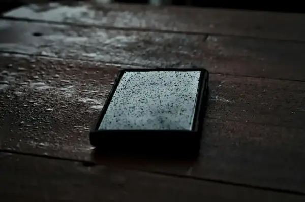 Telefon leży na mokrym drewnianym stole, pokryty kropelkami deszczu. Jego obudowa z certyfikatem IPX5 zapewnia mu ochronę przed wilgocią.