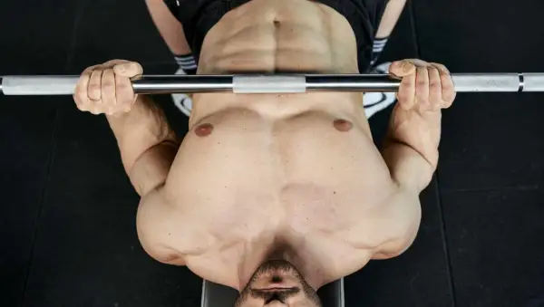 Uomo con pettorali definiti e addominali scolpiti solleva un bilanciere durante un allenamento in palestra.