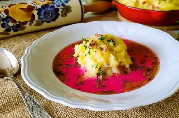 Pyszny barszcz czerwony zabielany, przepis babci. Na talerzu puree ziemniaczane z dodatkiem boczku i koperku, zanurzone w intensywnym, różowym barszczu.
