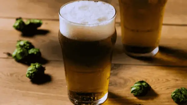 Bicchiere di birra dorata con schiuma bianca, circondato da luppoli verdi su un tavolo di legno. Perfetta per una dual IPA.