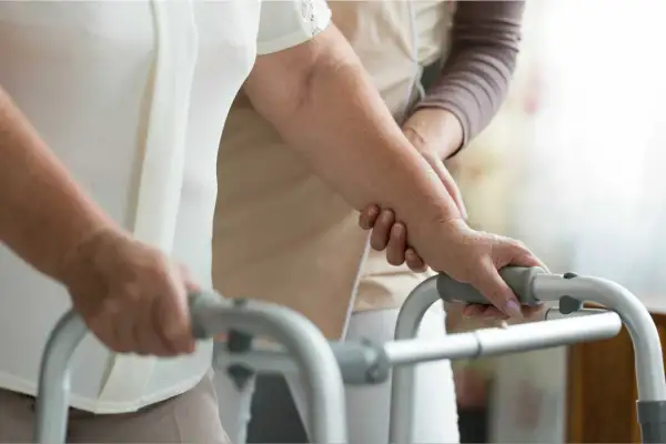 Una persona anziana, con l'aiuto di un'assistente, si appoggia a un deambulatore, mostrando la difficoltà nel riuscire a camminare all'improvviso.