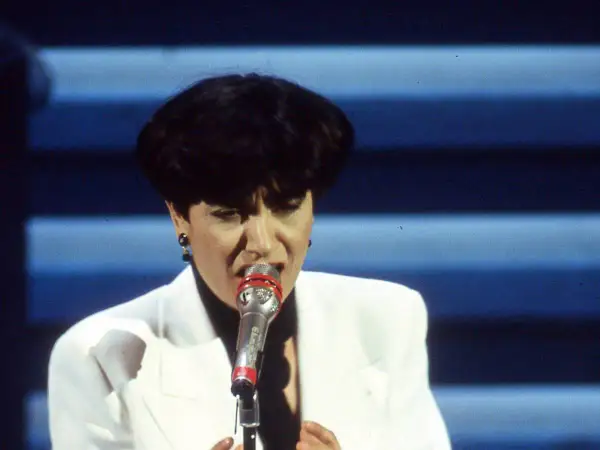 Mia Martini, icona della musica italiana, canta con passione sul palco, illuminata da luci soffuse.