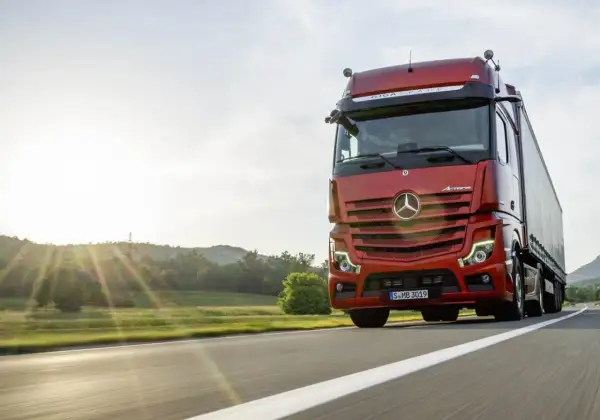 Un camion rouge vif roule sur une route ensoleillée, le soleil couchant illuminant le paysage. Ce camion sans rétroviseur semble prêt pour une longue route.