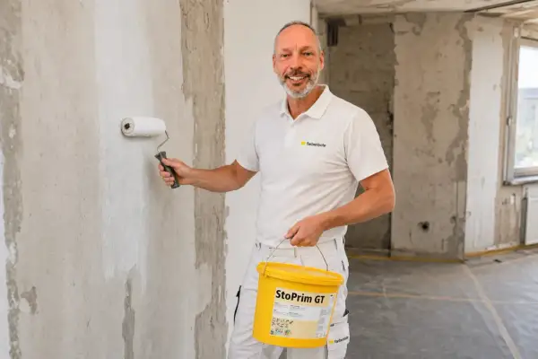 Maler grundiert mit einer Rolle die Wand. Er hält einen Eimer mit "StoPrim GT" in der Hand.
