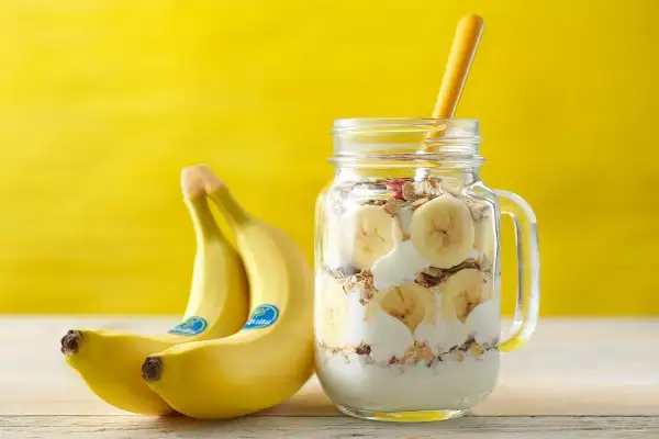 Un delizioso barattolo con yogurt, granola e fette di banana, perfetto per una banana a colazione. Accanto, due banane fresche su sfondo giallo.
