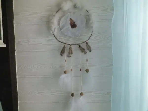 Un attrape rêve, ornement traditionnel, orné d'un loup hurlant, de plumes et de perles, suspendu devant un mur en bois clair.