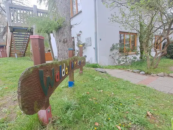 Holzschild "Waldorfkindergarten" vor einem Gebäude mit Spielturm. Mögliche Nachteile: wenig Struktur, viel Freiheit.