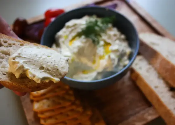 En bit bröd med krämig vispad fetaost och dill. I bakgrunden en skål med mer ostdipp och kex.