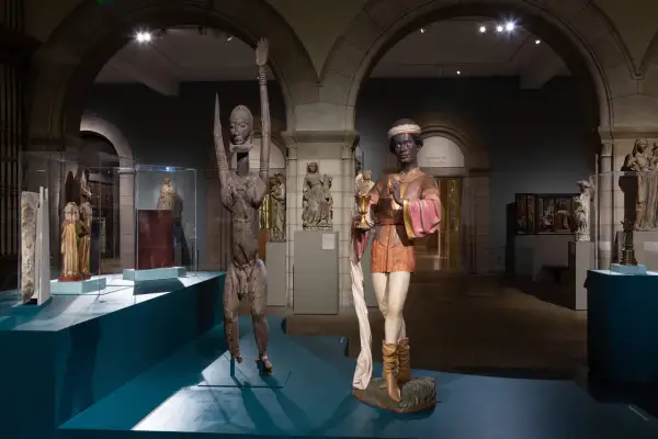 Two striking sculptures, one a wooden figure with raised arms, the other a painted figure holding a chalice, are displayed in a museum gallery, showcasing medieval African art.