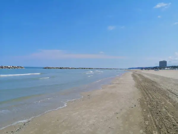 Spiaggia del lido di Fermo con mare calmo, frangiflutti in pietra e un palazzo sullo sfondo.