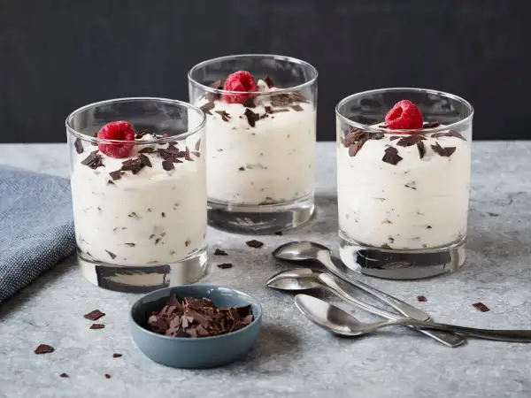Drei Gläser mit cremigem Dessert, garniert mit Himbeeren und Schokoladenstückchen. Ein Schälchen mit Schokostückchen und Löffel daneben. Ein leckeres Herrencreme Rezept zum Nachmachen.