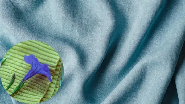 A blue tencel fabric drapes softly, hinting at its smooth, natural feel. A purple flower rests on a textured yellow surface.