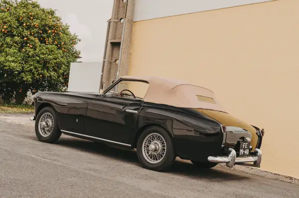 Svart, klassisk brittisk sportbil med beige sufflett parkerad vid en gul vägg, bredvid ett apelsinträd.