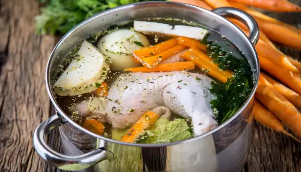 Pentola con brodo di pollo ricetta della nonna: pollo, carote, sedano, cipolla e prezzemolo in cottura su un tavolo di legno.