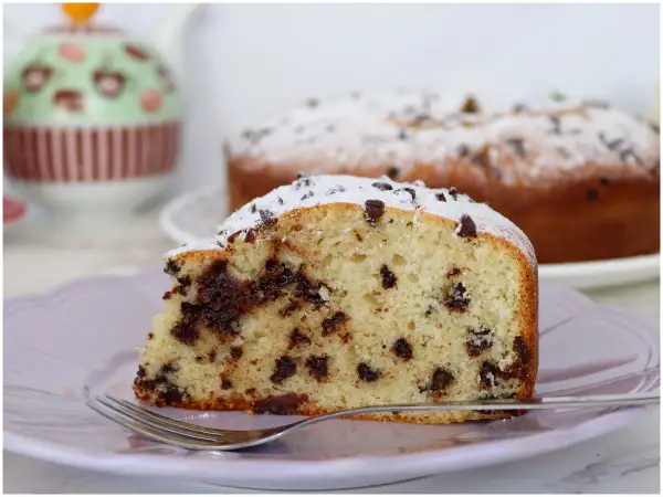 Una fetta di torta soffice con tante gocce di cioccolato, cosparsa di zucchero a velo, su un piatto lilla con una forchetta.