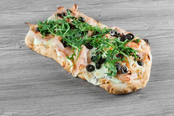 Pinsa co to? Podłużna, chrupiąca baza z szynką, mozzarellą, rukolą i oliwkami. Pyszna alternatywa dla pizzy.