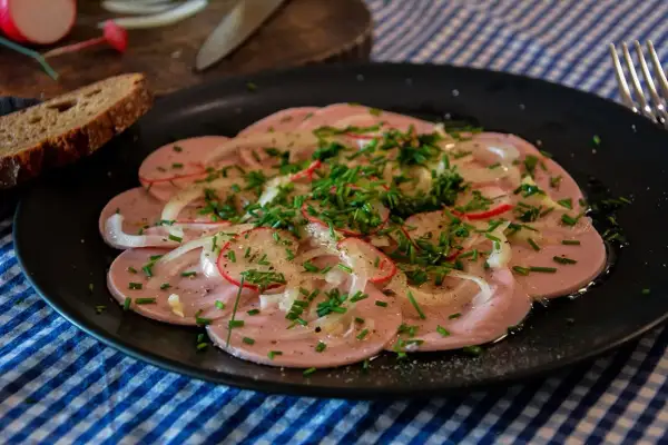 Saure Wurst mit Zwiebeln, Radieschen und Schnittlauch auf schwarzem Teller, dazu Brot.