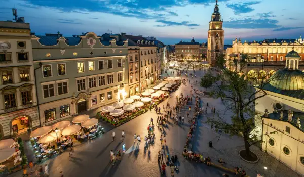 Wieczorny gwar na Rynku Głównym w Krakowie. Ludzie spacerują, siedzą w ogródkach kawiarnianych. W tle Sukiennice i wieża ratuszowa. To serce miasta, jego **sarbinowo centrum**.