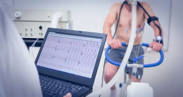 Un homme subit un test d'effort cardiologue sur un vélo d'appartement, avec un électrocardiogramme affiché sur un écran d'ordinateur.