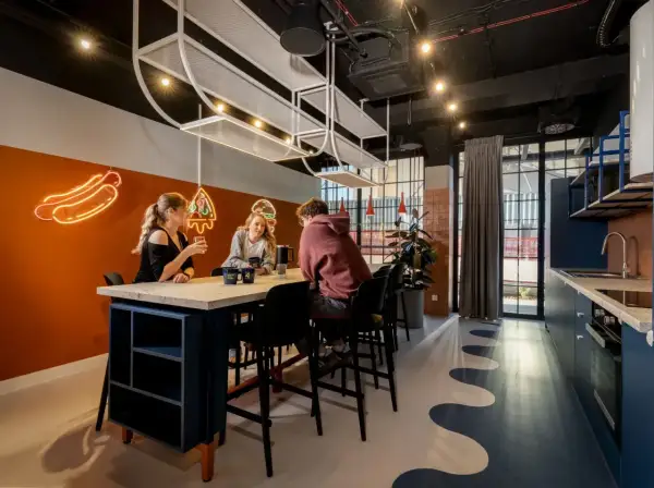 Studenci przy stole, rozmawiają i piją napoje. W tle neonowe hot dogi i pizza. To miejsce sprzyja nauce i relaksowi.