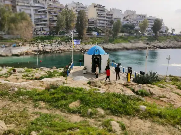Mała kapliczka nad wodą, z grecką flagą, na tle budynków. Widok na pireus plaża.