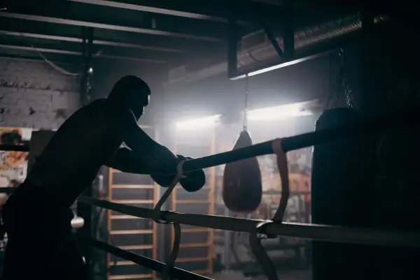 A boxer leans on the ropes in a dimly lit gym, contemplating how to get into boxing matches. Punching bags hang in the background.