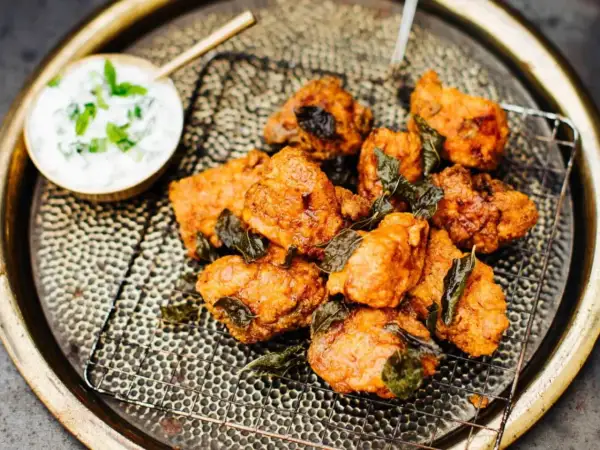 Knusprige chicken pakora, garniert mit frischen Kräutern, auf einem Gitterrost serviert. Daneben eine Schale mit Joghurt-Dip.