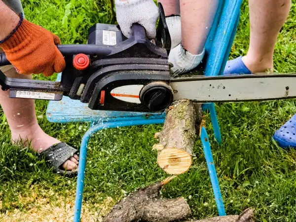 Osoba w pomarańczowej rękawicy używa piły łańcuchowej do cięcia drewna na prowizorycznym stojaku. To prosty sposób, jak zrobić stojak do cięcia drewna.