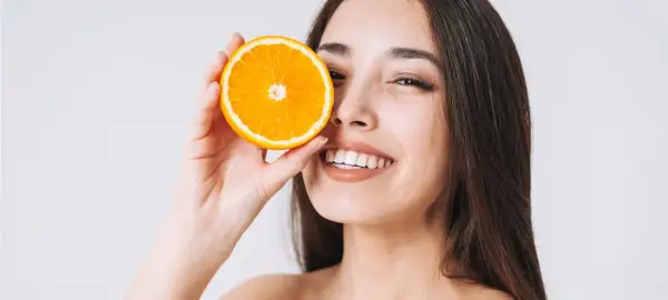 Lächelnde Frau hält Orangenscheibe vor ihr Auge. Vitamin C kann bei Rosacea helfen, die Haut zu beruhigen.