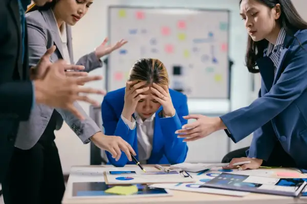Giovane donna d'affari stressata, con le mani sulla testa, mentre colleghi la circondano con gesti agitati. Lo stress lavoro correlato è un rischio da valutare.