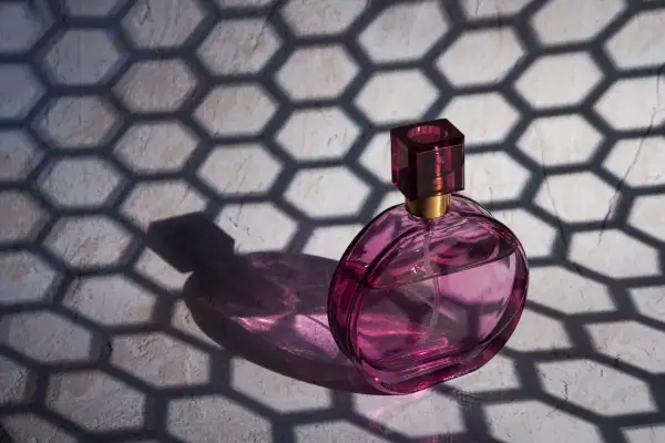 Różowa buteleczka perfum Dior J'adore, oświetlona słońcem, rzuca cień o geometrycznym wzorze plastra miodu.